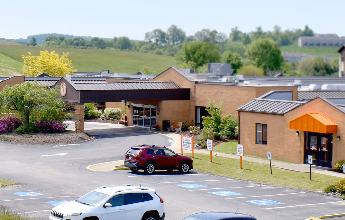 Exterior view of William Penn Care Center showing a single-story brick building with a covered entrance, surrounded by landscaped greenery and a parking lot with several cars parked, including handicap parking spaces.
