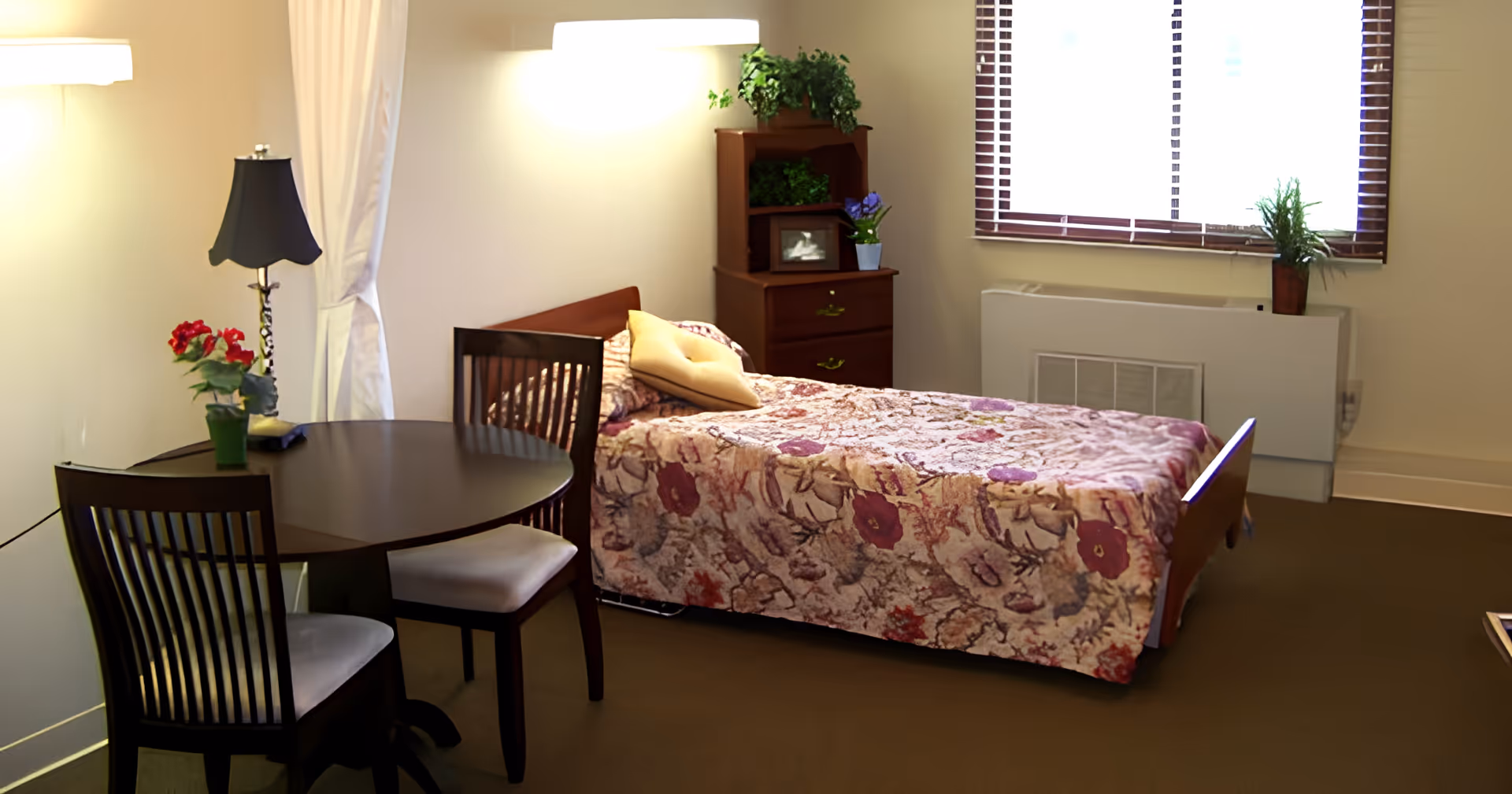 A cozy bedroom with a single bed covered in a floral patterned bedspread, a yellow pillow, a wooden nightstand with plants and a framed photo, a round wooden table with two chairs, a black lamp, and a window with blinds letting in natural light.