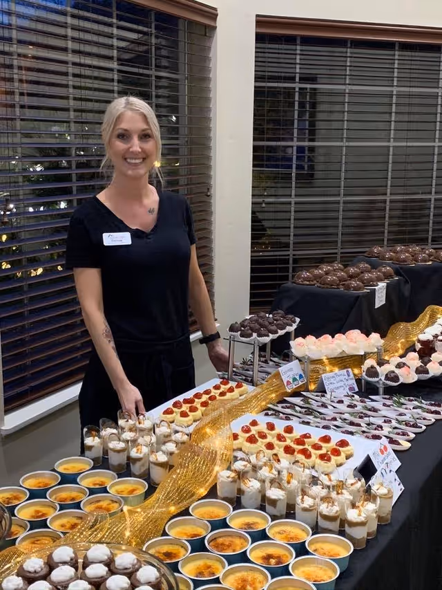 A smiling woman with blonde hair tied back, wearing a black shirt and a name tag, stands behind a table filled with an assortment of desserts including crème brûlée, mini cheesecakes topped with cherry, chocolate truffles, and layered parfaits. The setting appears to be indoors with large windows and blinds in the background.