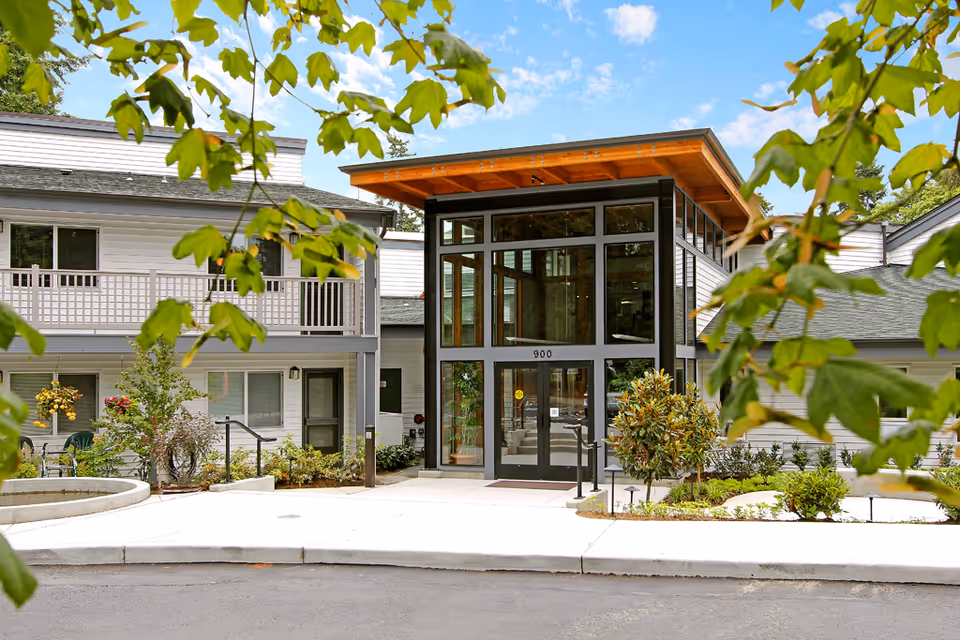 Entrance of a modern senior living building with a glass-walled lobby, balconies, and landscaping seen through tree leaves.