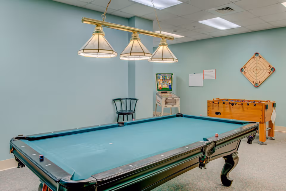 A recreational room with a pool table in the foreground, a foosball table, and a vintage pinball machine against a light blue wall. There is a black chair in the corner and a wooden game board mounted on the wall. Three pendant lights hang above the pool table.