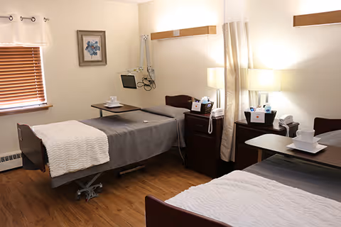 A clean and well-lit room with two hospital-style beds, each with gray bedding and white blankets. Each bed has an overbed table with a white cup and saucer. There are two bedside tables with lamps, a telephone, and some medical supplies. A window with wooden blinds and a framed picture are visible on the wall.