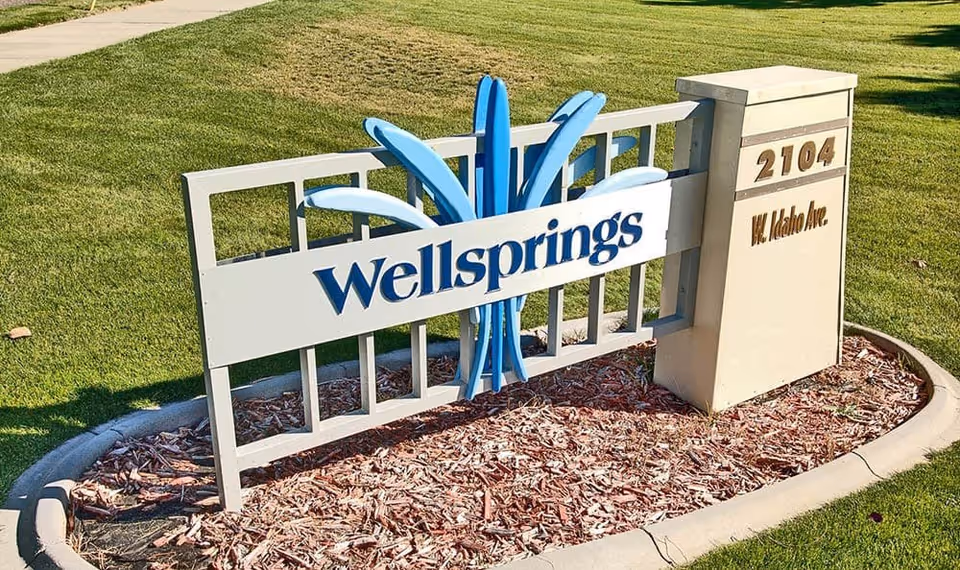 Outdoor sign for Wellsprings Assisted Living located at 2104 W. Idaho Ave, set in a landscaped area with grass and mulch.