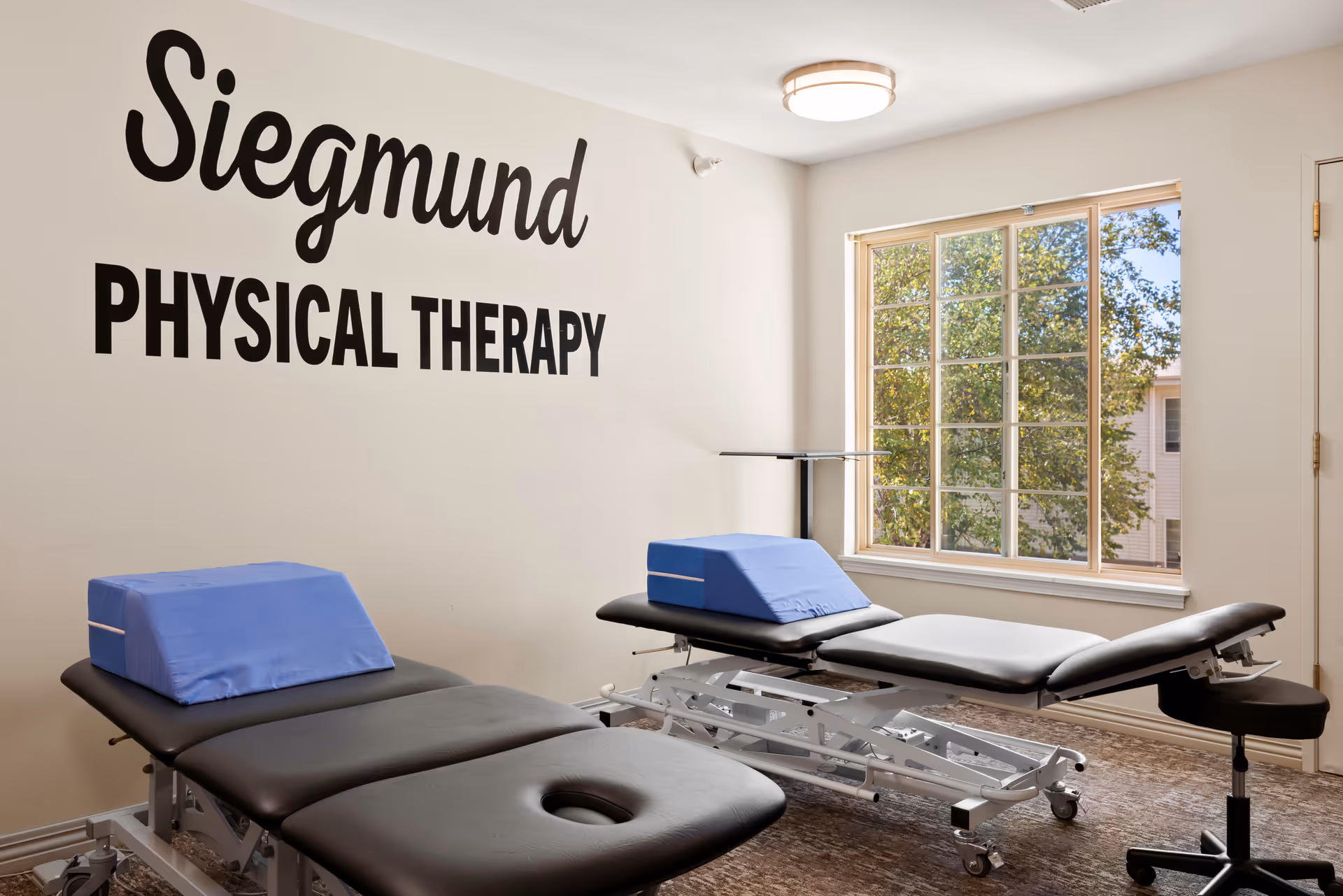 A physical therapy room with two black treatment tables, each with a blue wedge cushion. The room has a large window letting in natural light and a wall sign that reads 'Siegmund PHYSICAL THERAPY'.