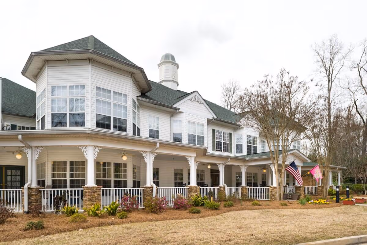 Front exterior of a two-story senior living building with a wraparound porch, columns, landscaping, and flags.
