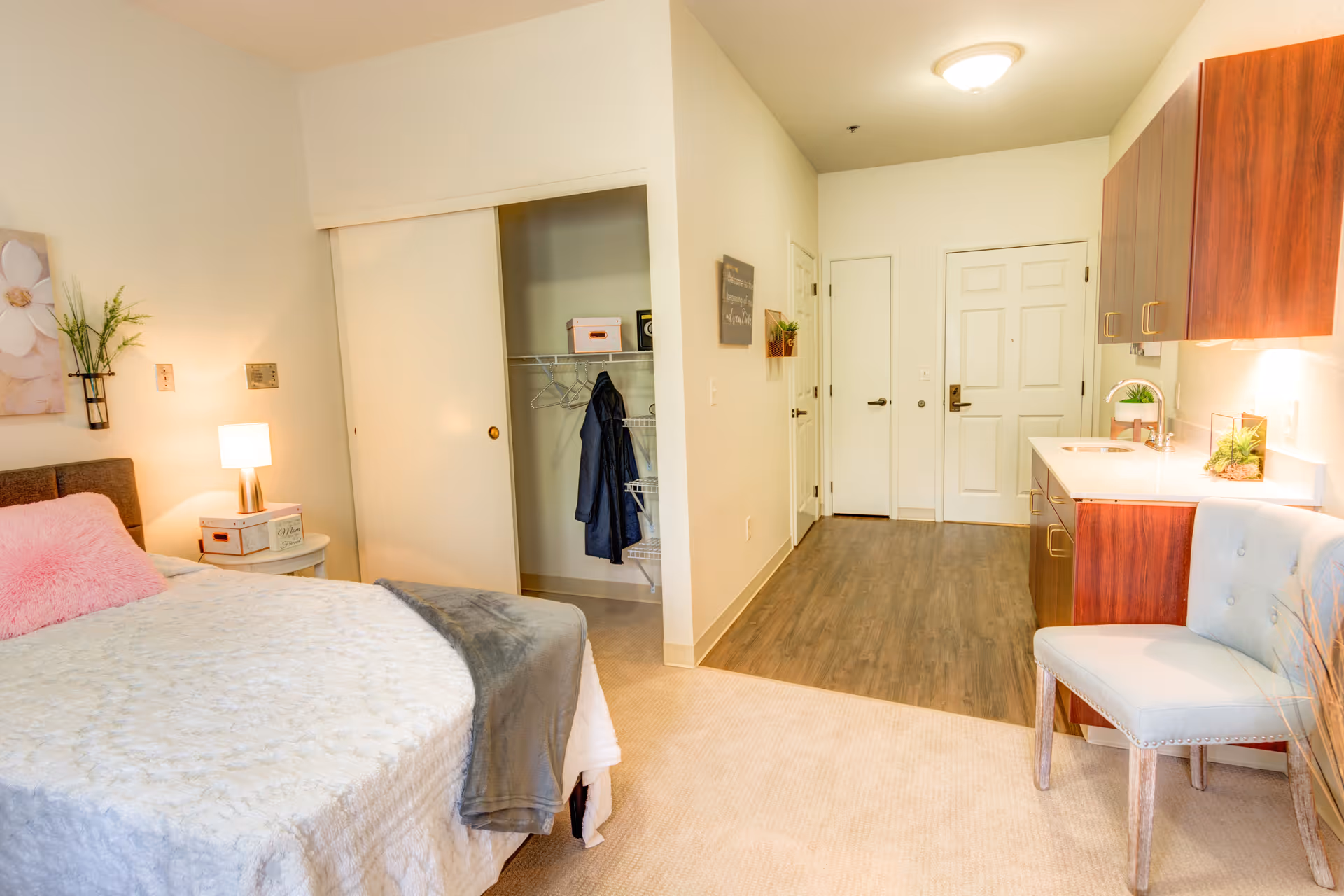 A cozy senior living studio apartment featuring a bed with a pink pillow and gray blanket, a small nightstand with a lamp and decorative items, an open closet with a hanging coat, a kitchenette with wooden cabinets and a sink, a light blue upholstered chair, and a hallway leading to the entrance door.