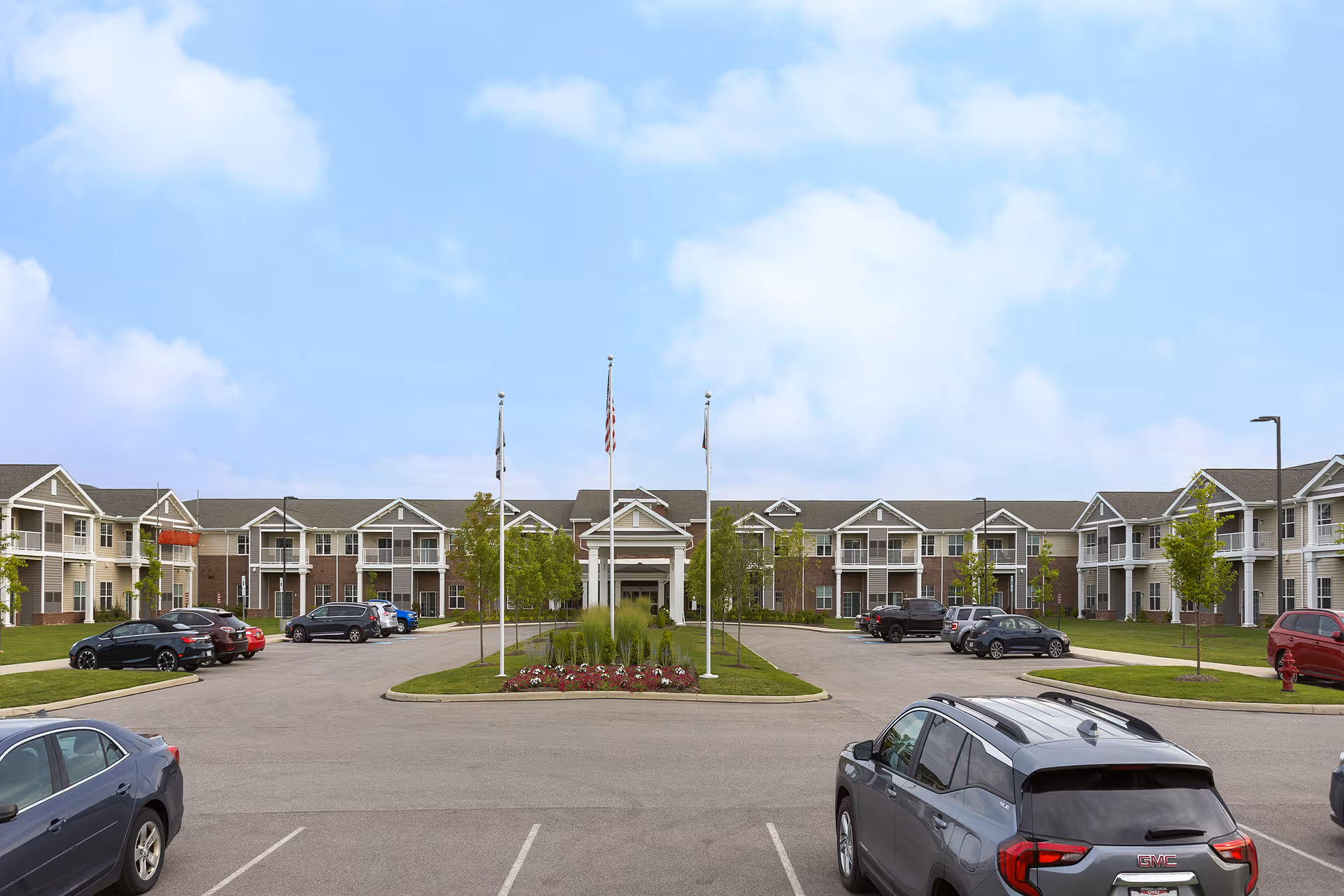 Front exterior view of StoryPoint Medina senior living facility with a parking lot in the foreground, several parked cars, a landscaped island with three flagpoles, and a two-story building with balconies under a partly cloudy sky.
