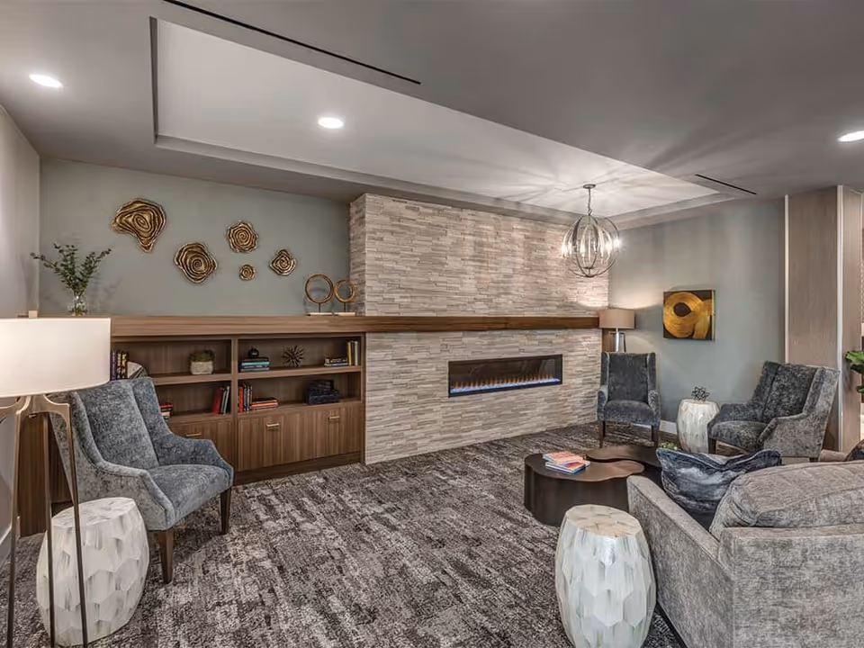 A modern communal lounge with gray upholstered chairs and sofas arranged around a coffee table in front of a linear fireplace set into a stone accent wall.