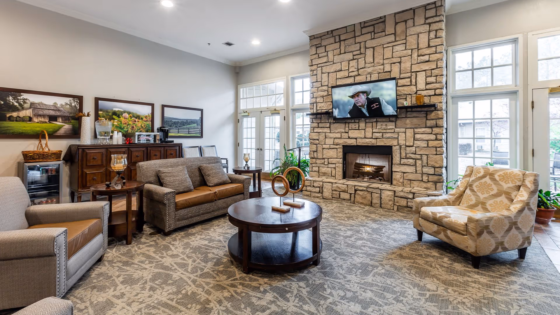 A cozy living room area in Hearthstone Assisted Living featuring a stone fireplace with a mounted flat-screen TV above it. The room has a patterned carpet, a round wooden coffee table with decorative items, a beige patterned armchair, a gray armchair, and a gray sofa with brown cushions. Large windows and glass doors allow natural light to fill the space. On the left side, there is a wooden cabinet with framed landscape pictures above it and a small beverage fridge underneath.