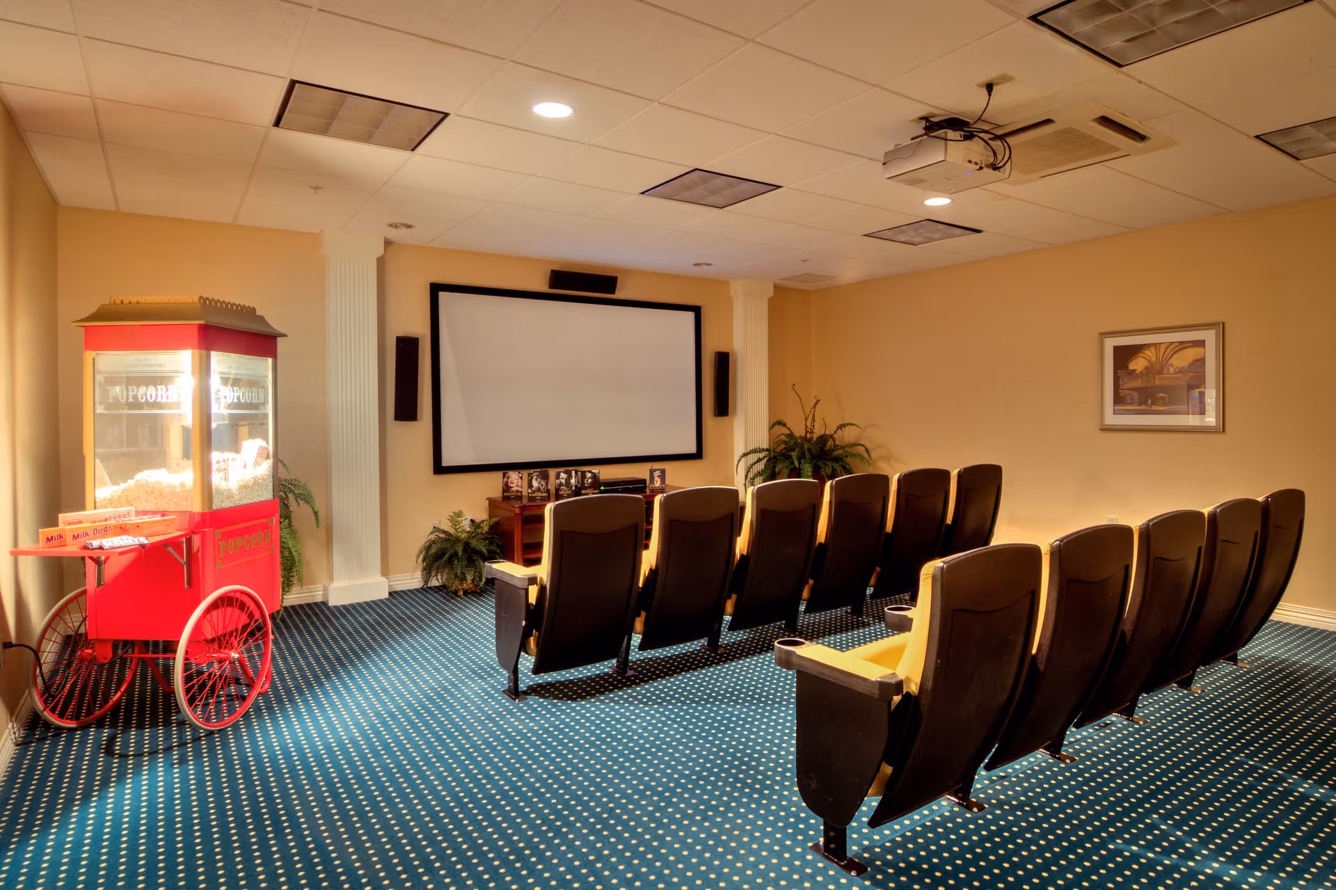 A small movie theater room with two rows of yellow and black theater seats facing a large blank projection screen. To the left, there is a red popcorn machine filled with popcorn and some candy boxes on a small shelf. The room has beige walls, a blue carpet with white dots, and some green plants near the screen. A projector is mounted on the ceiling.