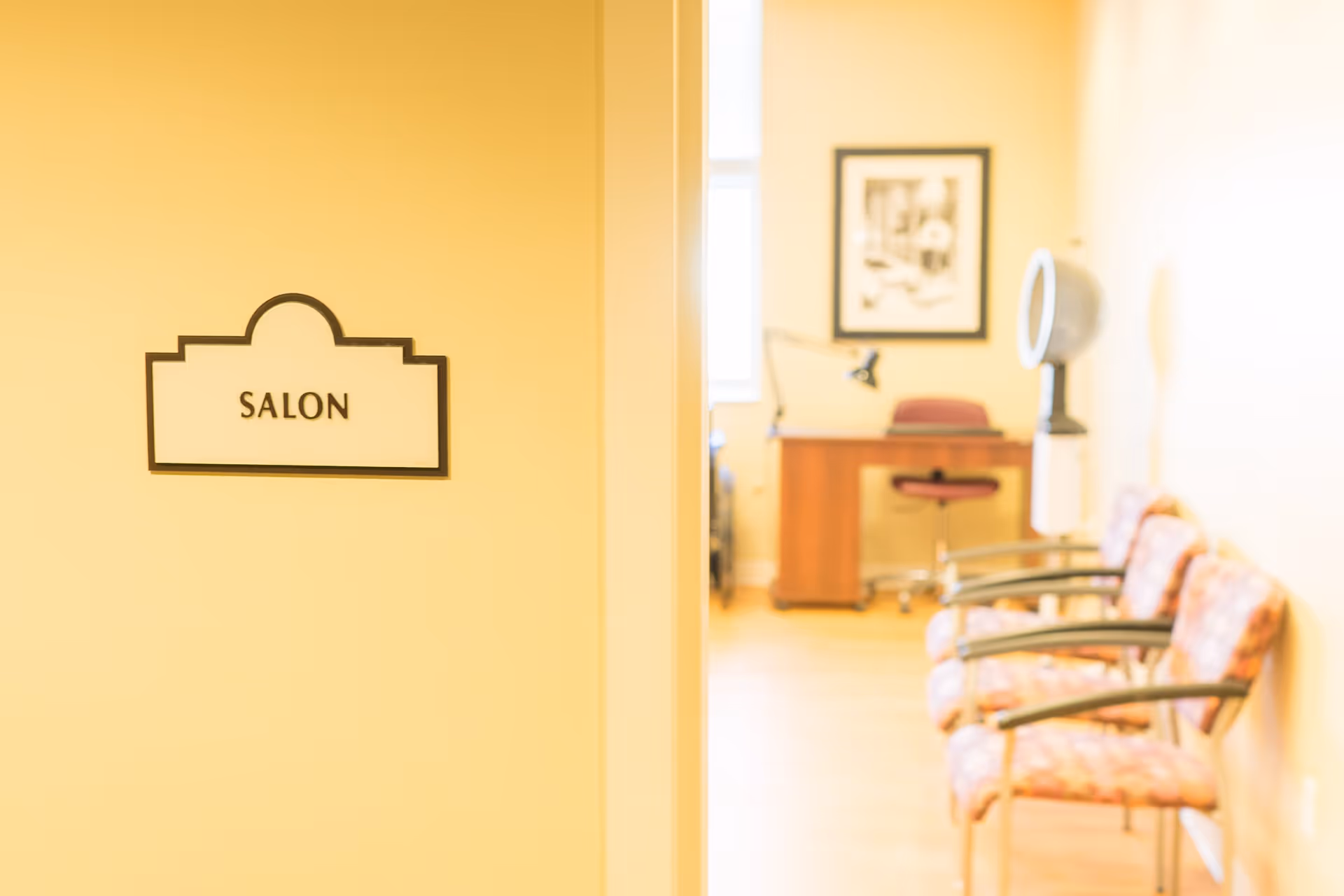 Interior view of a salon in a senior living facility with a sign labeled 'SALON' on a yellow wall. Inside the room, there are three patterned chairs lined up against the right wall, a wooden desk with a chair, a lamp, and a framed picture on the far wall. A hair dryer is also visible near the chairs.
