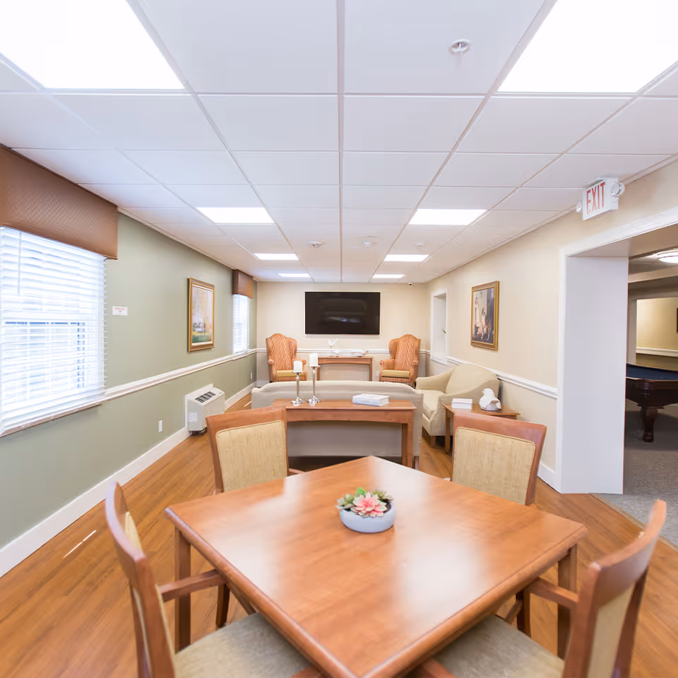 A bright and spacious senior living common area with a wooden table and four chairs in the foreground, a seating area with armchairs and a sofa, a flat-screen TV mounted on the wall, framed artwork, and large windows with blinds allowing natural light.