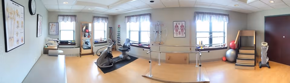 A rehabilitation room with exercise equipment including a recumbent bike, parallel bars for walking practice, therapy stairs, exercise balls, anatomical charts on the walls, and large windows with curtains letting in natural light.