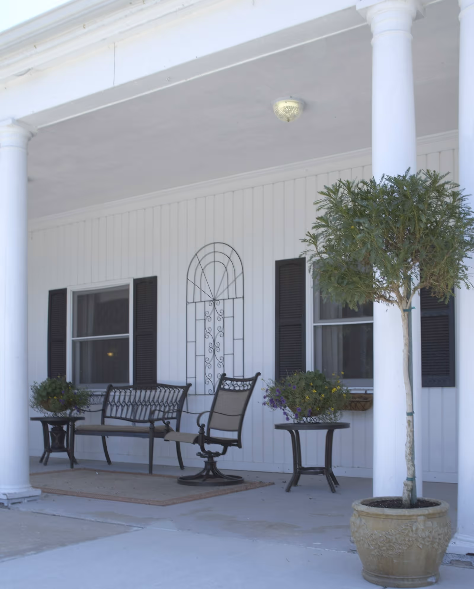 Covered porch area with white columns, a bench, a swivel chair, two side tables with flower pots, and a potted tree in a large decorative planter. The porch has white siding and two windows with black shutters.