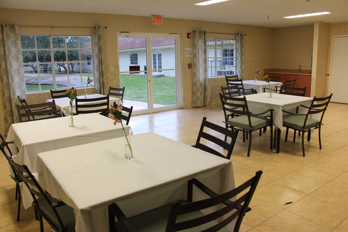 Dining room with several tables draped in white cloths and chairs, each table holding a single flower, and large windows and glass doors looking out to a courtyard.