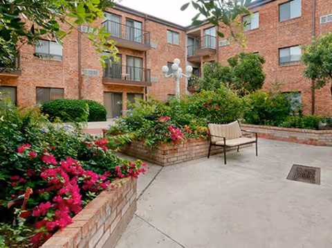 Outdoor courtyard area of a senior living facility with brick planters filled with green shrubs and pink flowers, a bench, and a multi-globe lamp post. The courtyard is surrounded by a three-story brick building with balconies and windows.