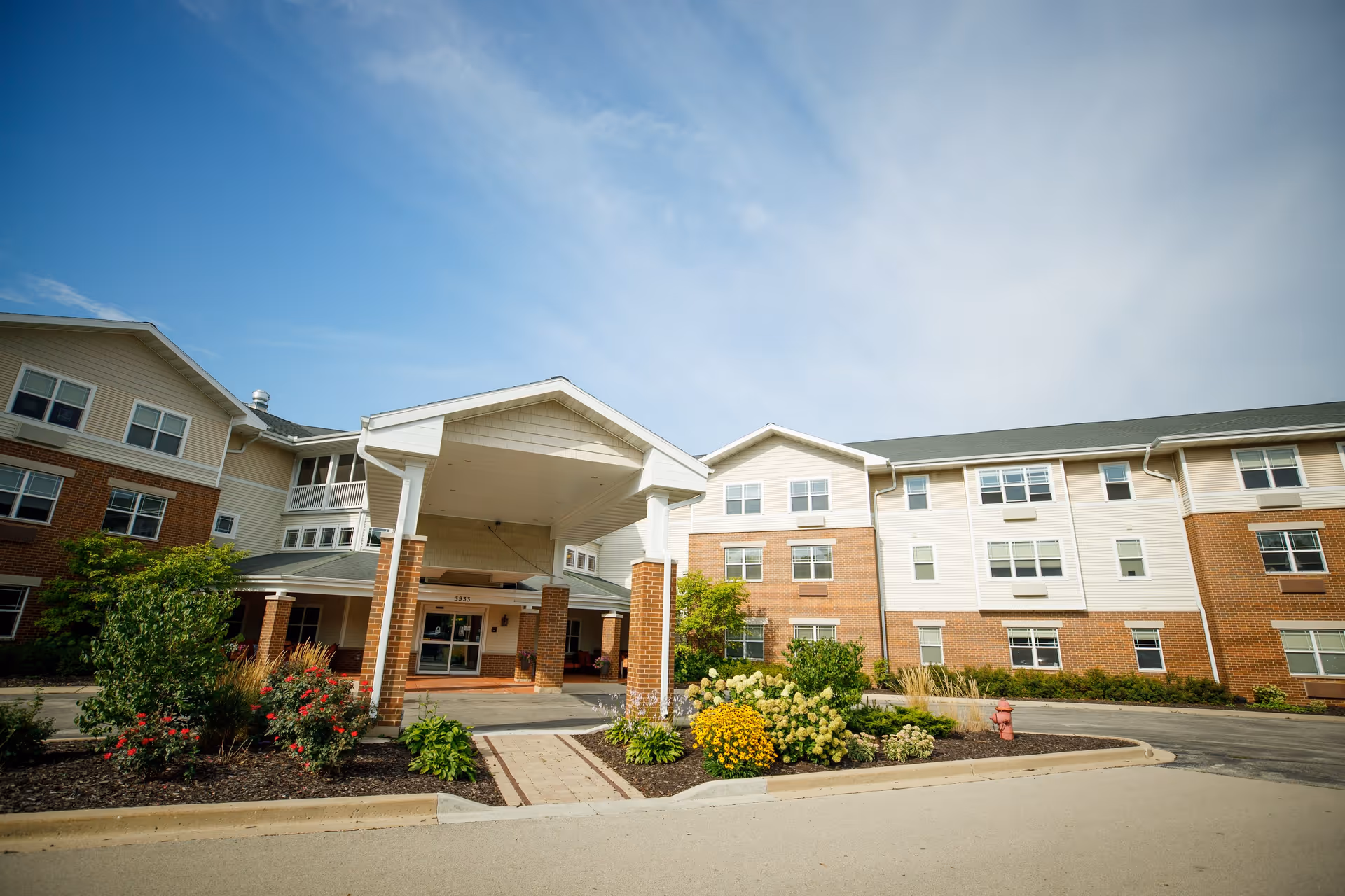 Exterior view of a senior living facility building with a covered entrance, landscaped garden beds with flowers and shrubs, and a clear blue sky.