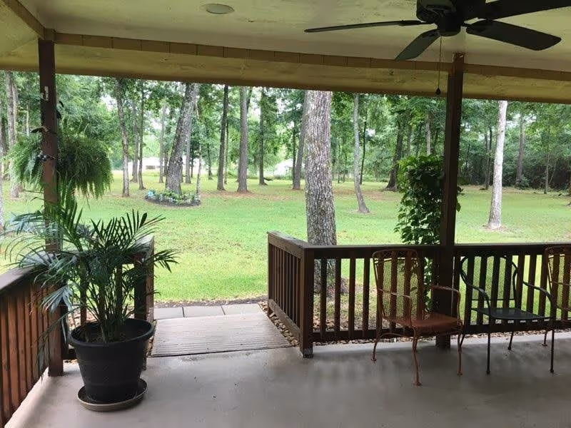 Covered patio area with wooden railing, a ceiling fan, potted plants, and metal chairs overlooking a grassy yard with trees.