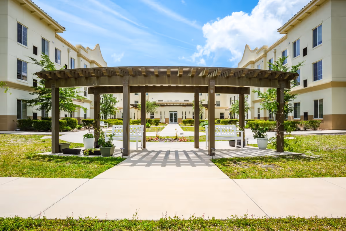 Outdoor courtyard area of a senior living facility with a wooden pergola structure, white benches, potted plants, and a pathway leading to a multi-story building under a blue sky with some clouds.