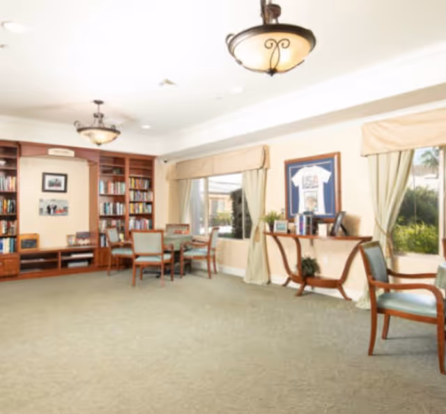 Bright common room with bookshelves, tables and chairs, framed artwork and large windows.