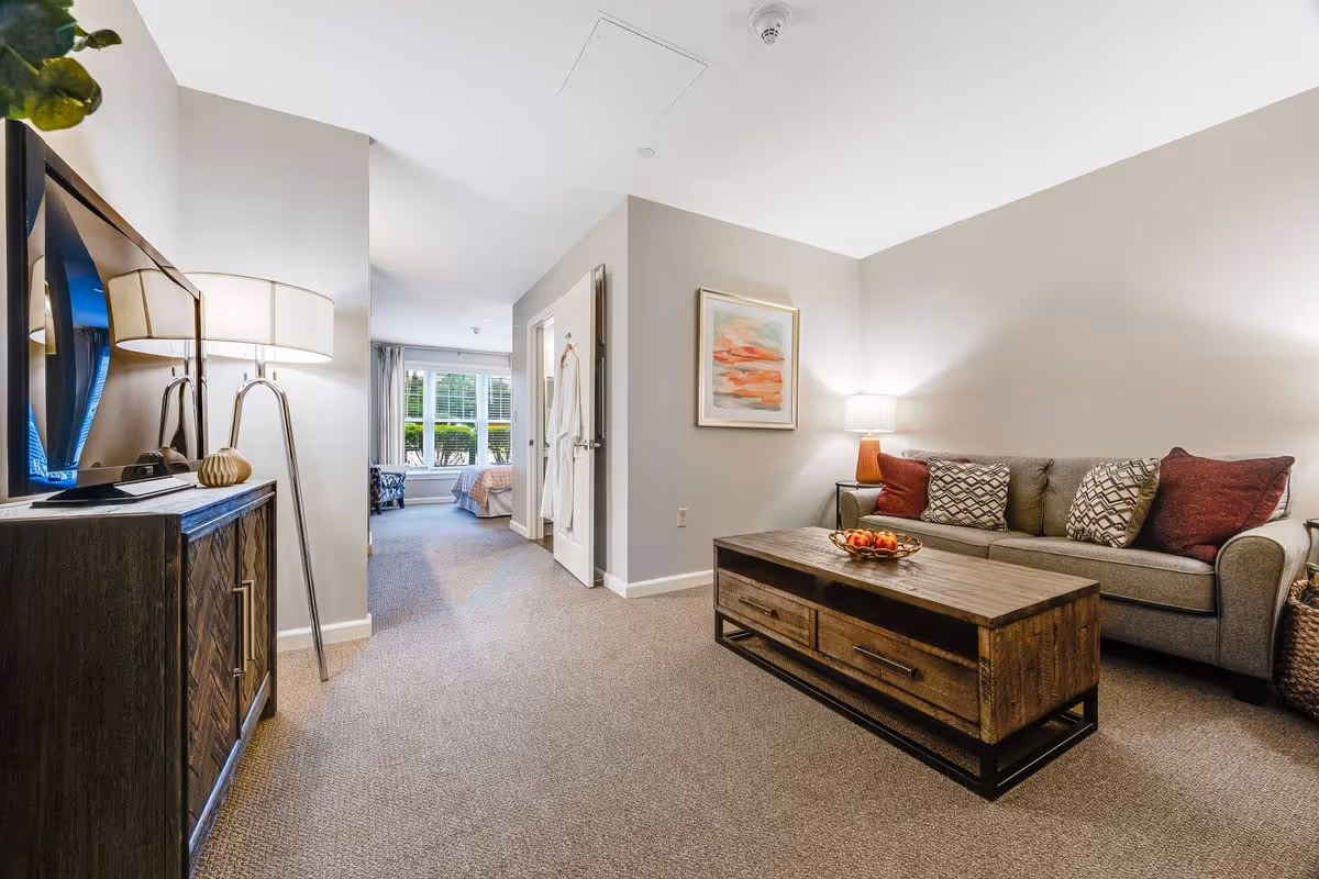 Bright furnished living room with a sofa and wooden coffee table facing a TV cabinet, leading down a hallway to a bedroom area.