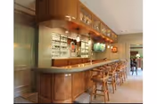 Interior bar/lounge area with a wooden counter, several bar stools, and shelving holding bottles and glassware.