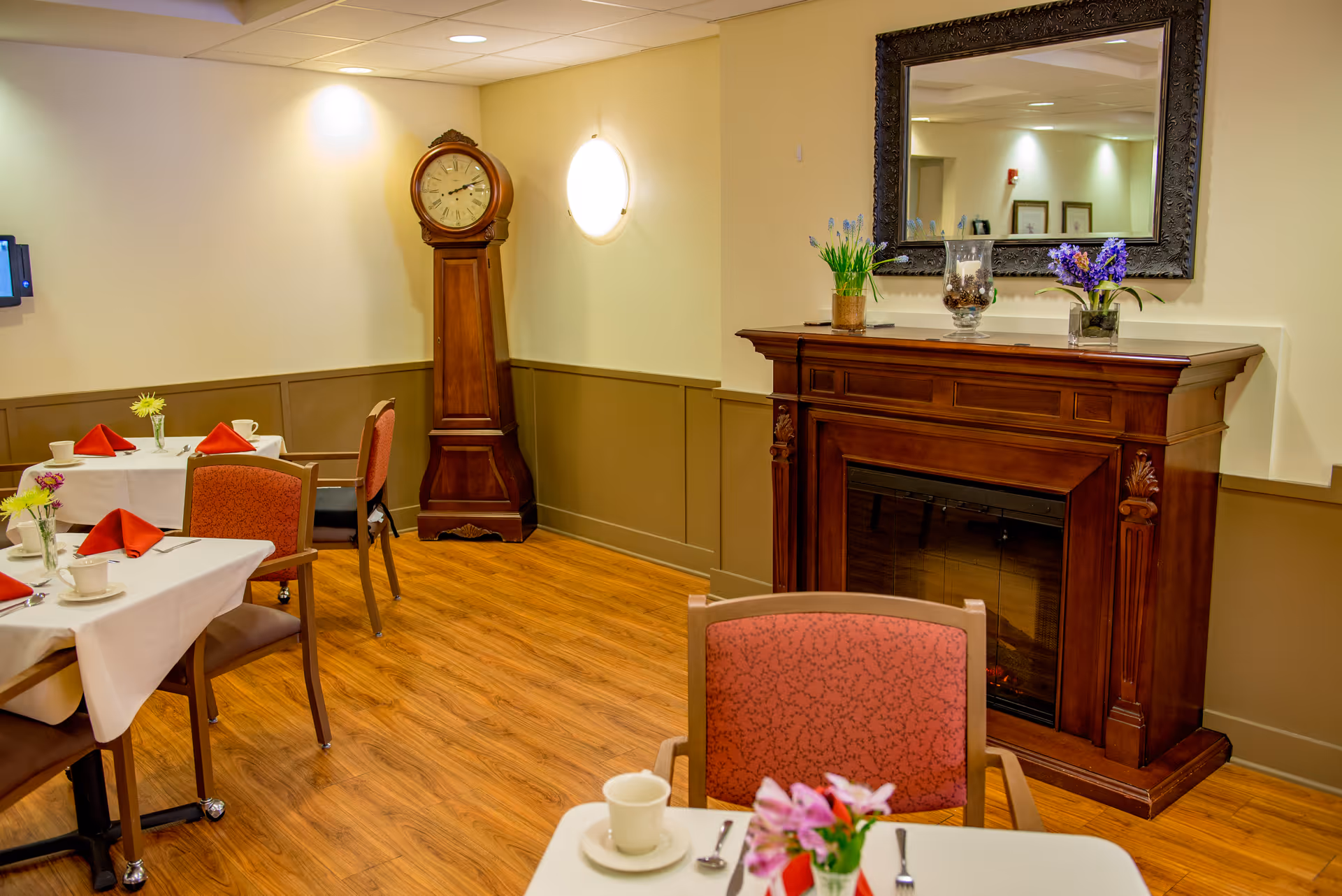 A cozy dining room with wooden flooring, tables set with white tablecloths, red napkins, cups, and small flower vases. There is a tall wooden grandfather clock in the corner and a wooden fireplace mantel with a large ornate mirror above it. The mantel is decorated with flower arrangements and a glass vase.