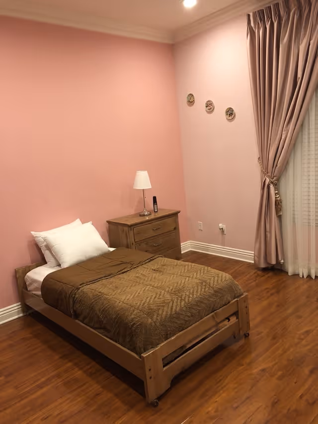 A simple bedroom with a single wooden bed covered with a brown blanket and white pillows. Next to the bed is a wooden nightstand with a small white lamp and a remote control. The room has pink walls, wooden flooring, three decorative plates on the wall, and a window with pink curtains tied back.