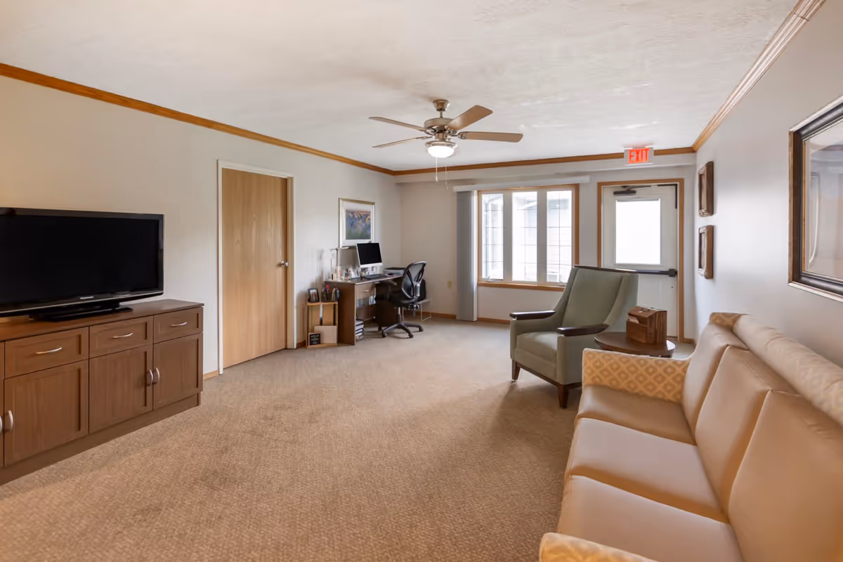 Spacious furnished common room with a sofa, armchair, television on a cabinet, a desk, and an exit door.