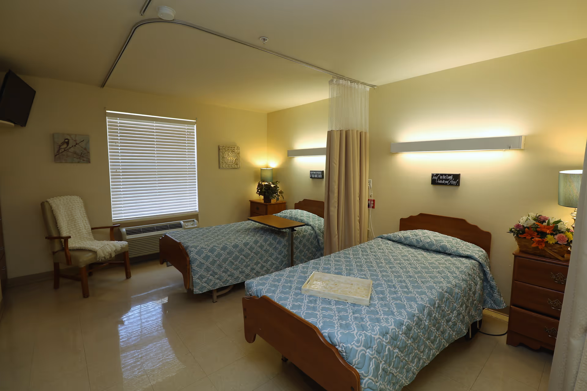 A shared bedroom in a senior living facility with two single beds separated by a privacy curtain. Each bed has a blue patterned bedspread. There is a wooden nightstand with a lamp and flowers next to one bed, and a chair with a white throw blanket near a window with closed blinds. The room has soft lighting and a clean, simple decor.