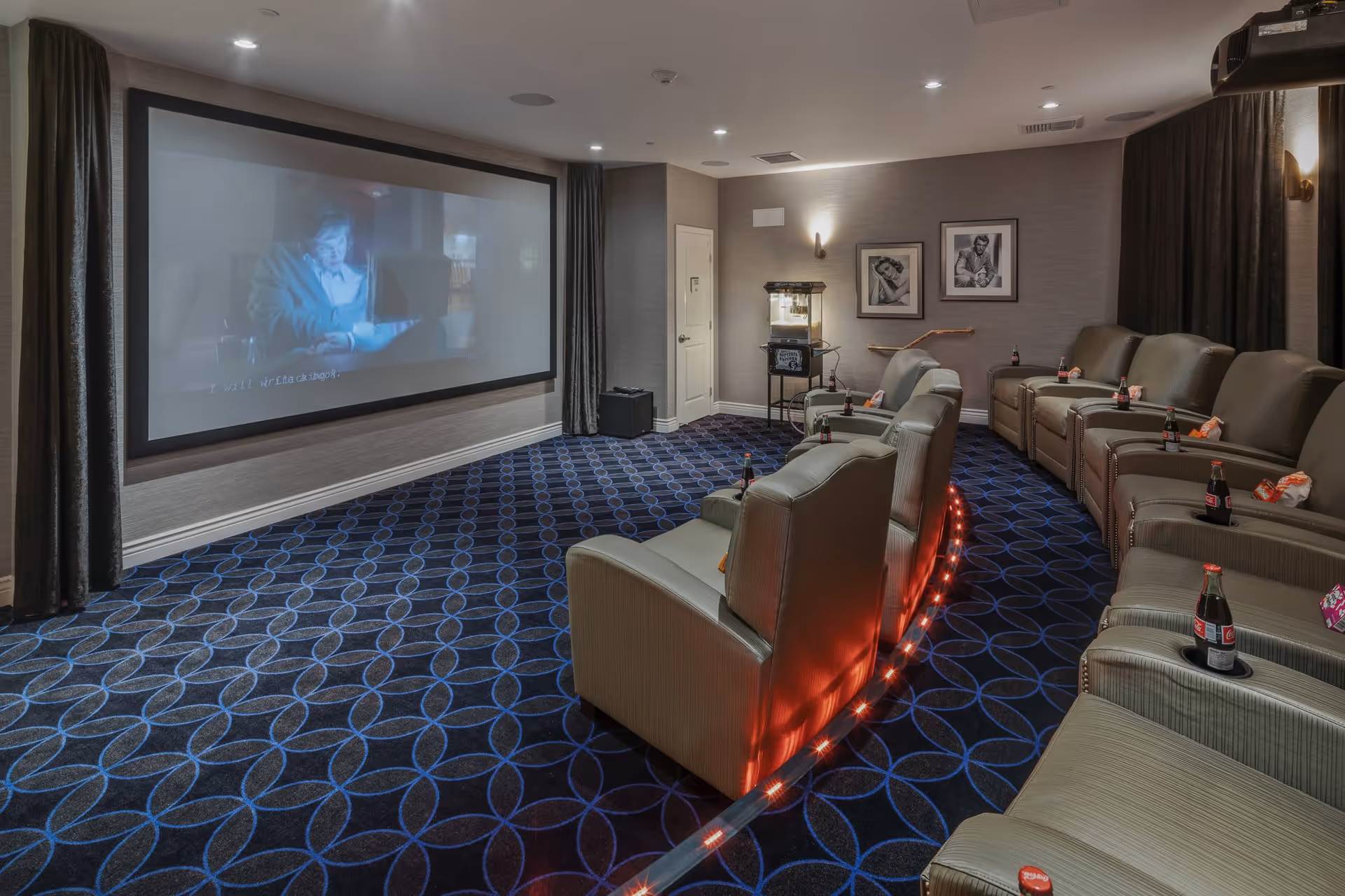 A cozy movie theater room with a large screen showing a film. The room features two rows of comfortable reclining chairs with cup holders, each holding a bottle of Coca-Cola and a bag of snacks. The carpet has a blue circular pattern, and the walls are decorated with framed black and white photos. There is a popcorn machine in the back corner of the room.