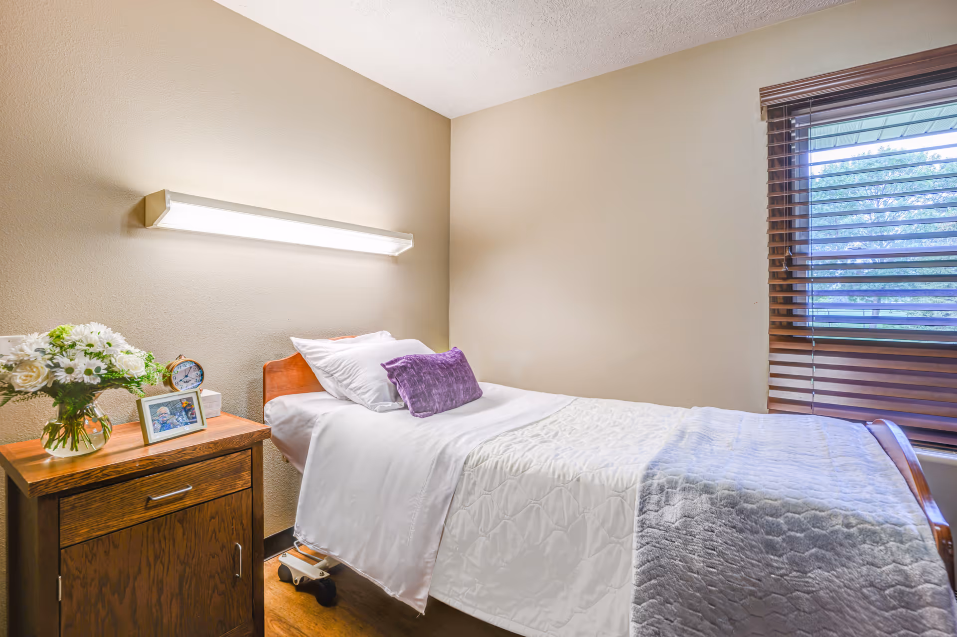 A small, neatly made single bed with white sheets, a white quilt, and a purple accent pillow in a room with beige walls. Next to the bed is a wooden nightstand with a vase of white flowers, a framed photo, and a clock. A window with wooden blinds is on the right side of the room, letting in natural light.