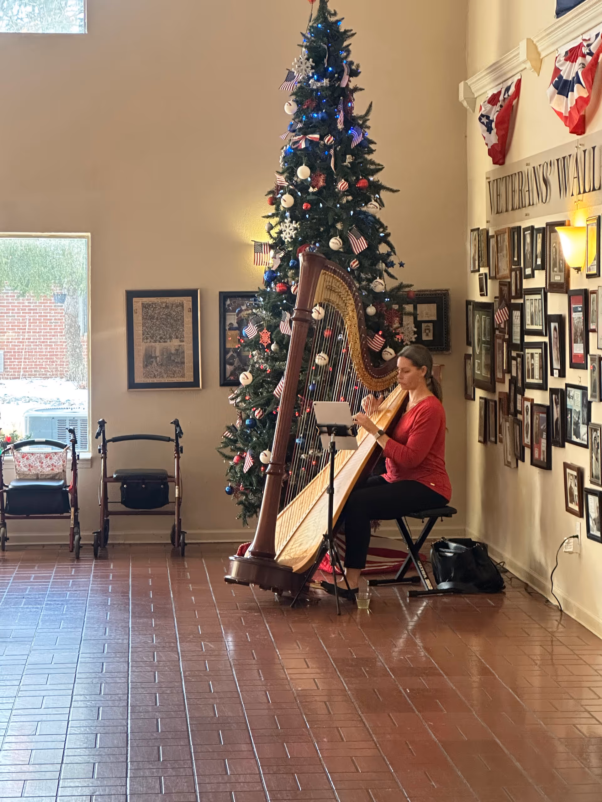 A musician plays a harp beside a decorated Christmas tree in a communal lobby area with framed photos on the wall.