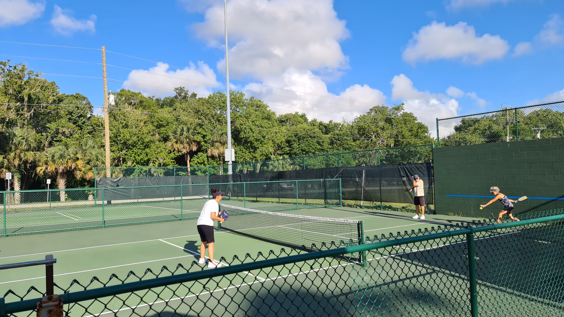 Three people playing pickleball on an outdoor court surrounded by green fencing, with trees and a blue sky with clouds in the background.