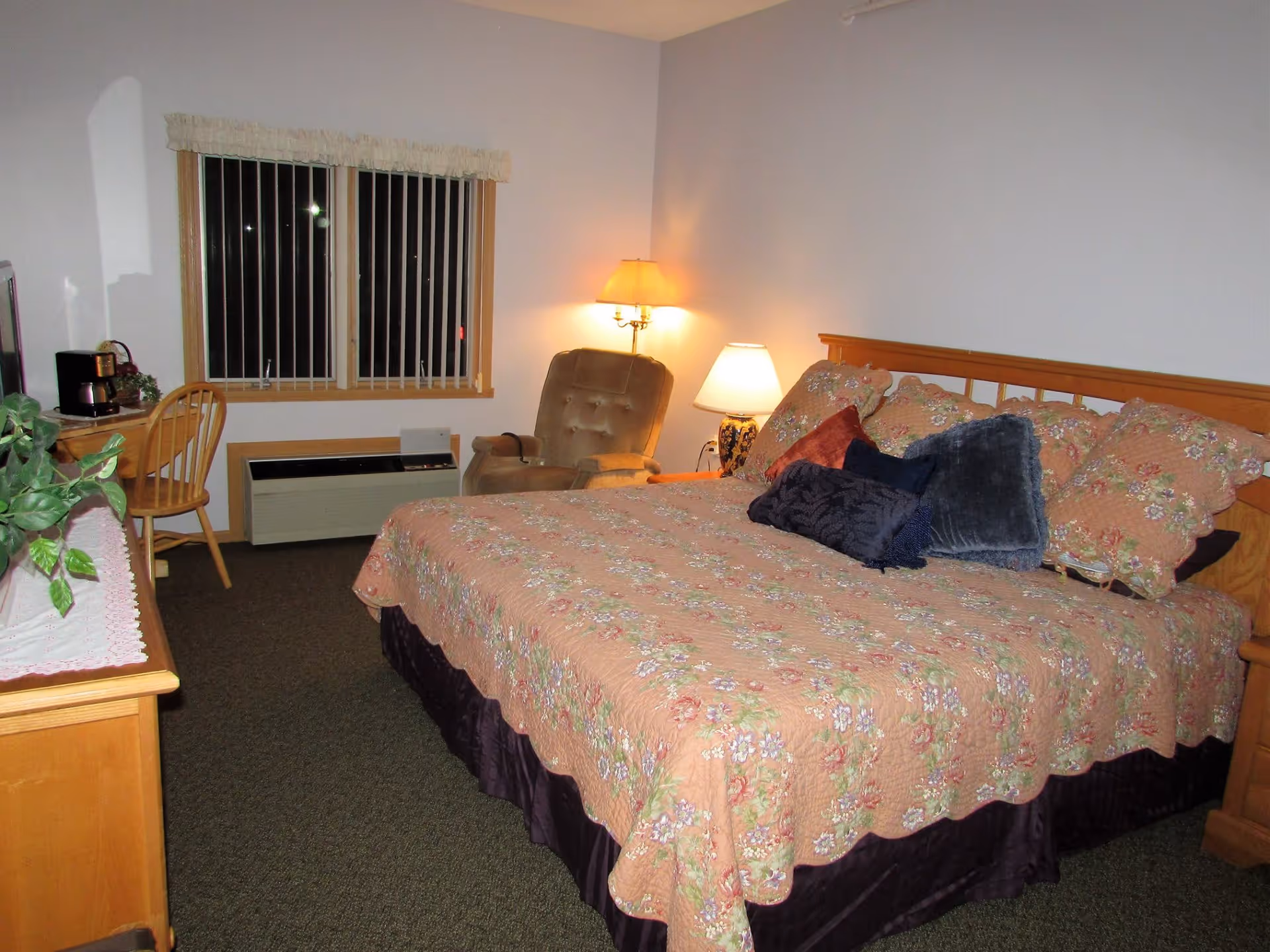 A cozy bedroom with a large bed covered in a floral quilt and multiple pillows. There is a cushioned armchair and two lamps providing warm light in the corner. A wooden desk with a chair, a coffee maker, and a plant are visible near a window with vertical blinds. The room has carpeted flooring and light-colored walls.