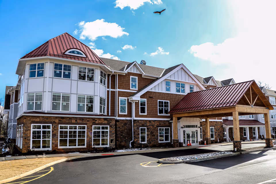 Front exterior of a multi-story senior living building with a covered porte-cochère entrance and red metal roofs under a blue sky.