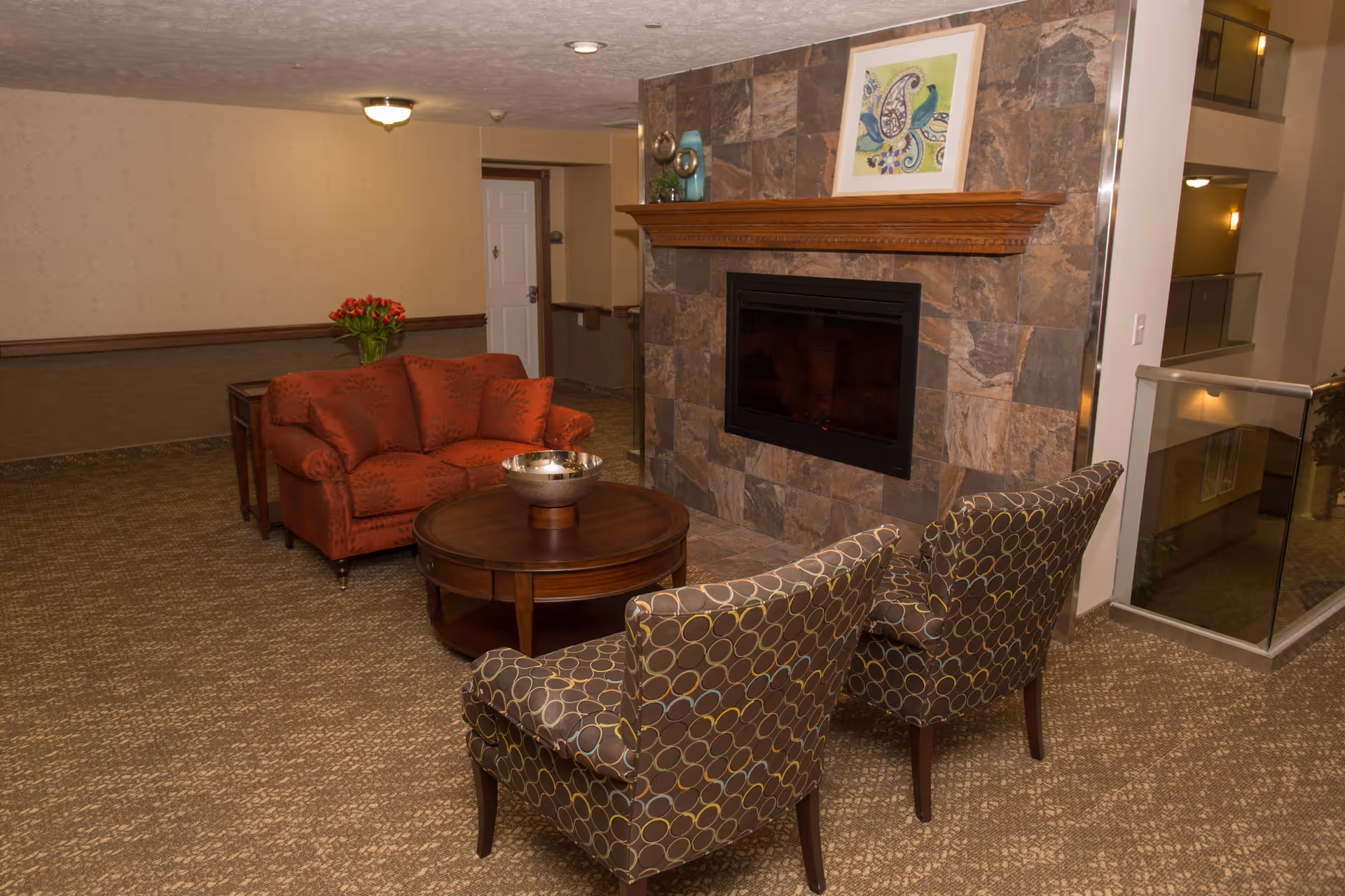 A cozy seating area in a retirement facility featuring a red loveseat, two patterned armchairs, a round wooden coffee table with a decorative bowl, and a stone fireplace with a wooden mantel. A framed artwork is displayed above the fireplace, and a vase with red flowers is on a side table behind the loveseat. The area is carpeted and adjacent to a hallway and an open atrium with glass railings.