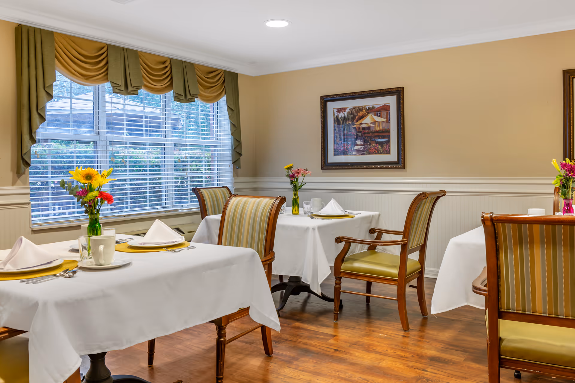 A dining room with tables covered in white tablecloths, each set with plates, napkins, cups, and silverware. The room has wooden chairs with green and gold striped cushions, a large window with green and gold drapes, and a framed painting on the beige wall. Fresh flowers in vases are placed on the tables.