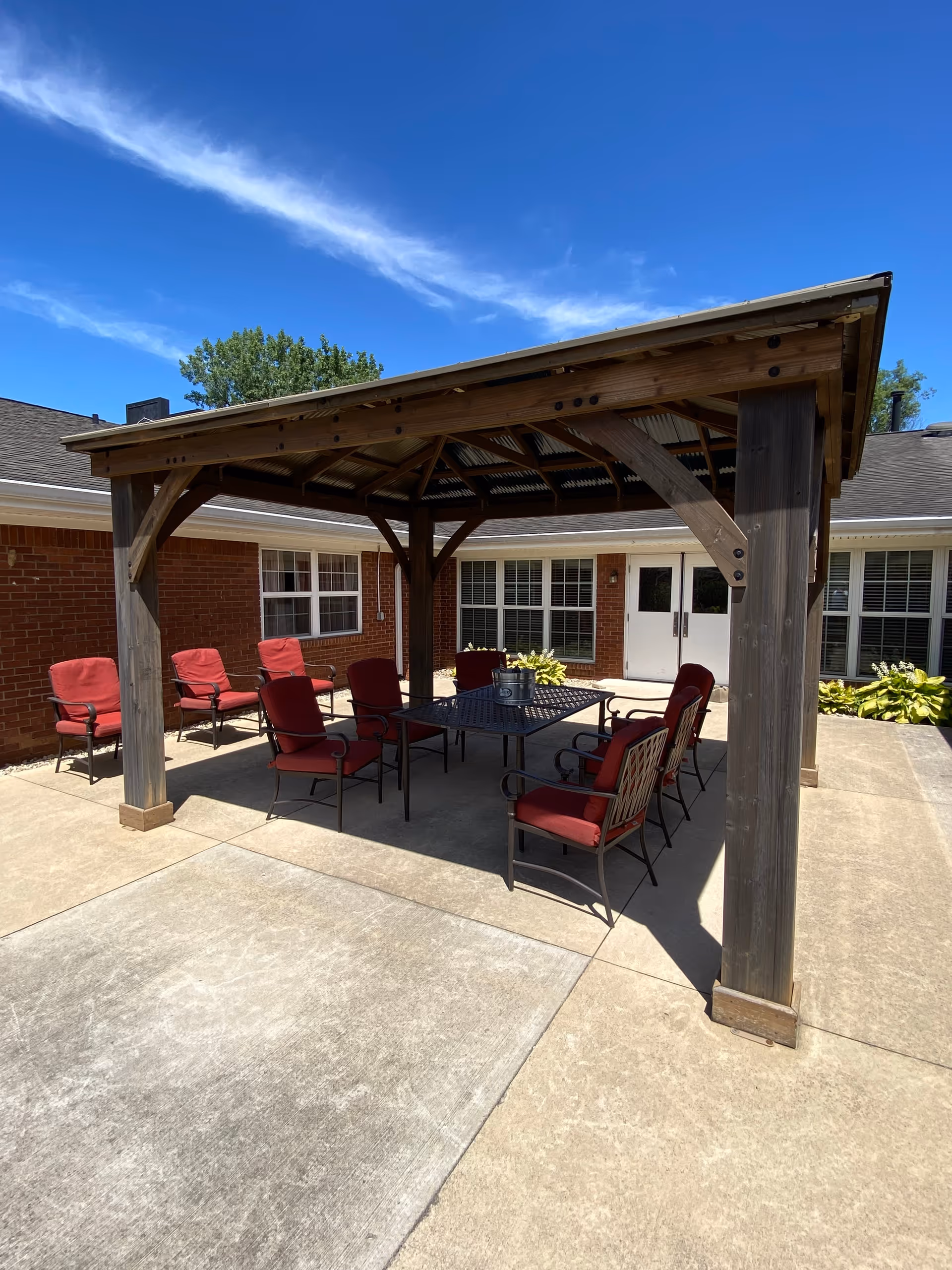 Outdoor patio area at Brickyard Healthcare - Brandywine Care Center featuring a wooden pergola with a metal roof, surrounded by red cushioned chairs and a black metal table. The patio is paved with concrete and the building's brick exterior and windows are visible in the background under a clear blue sky.