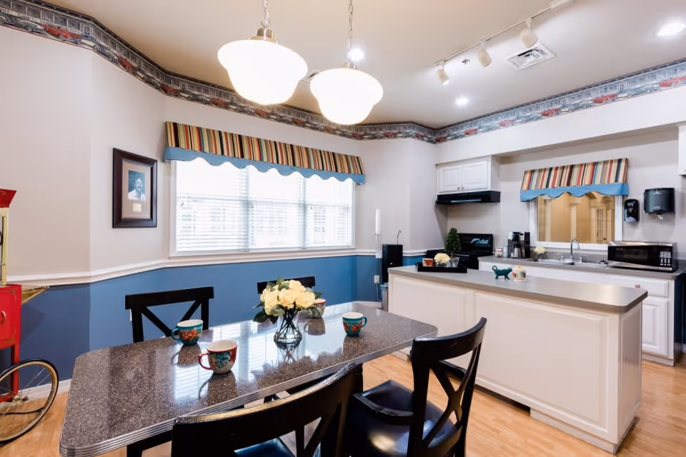 A bright kitchen and dining area in a senior living facility with a granite dining table set with four chairs and decorated with a vase of white flowers and colorful mugs. The kitchen features white cabinetry, a countertop island, a microwave, a coffee maker, and a window with striped valances. The walls are painted blue and white with a decorative border near the ceiling.