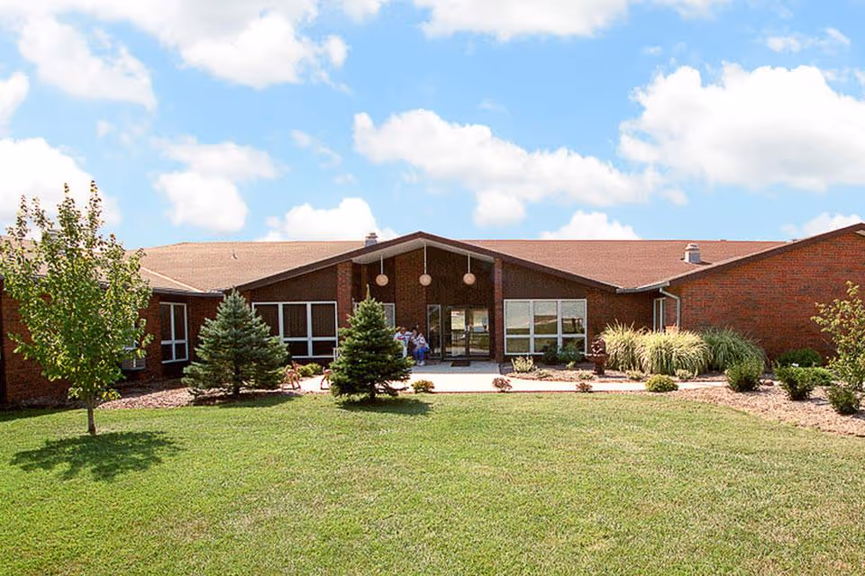 Front exterior view of a single-story brick building with a peaked roof, surrounded by a well-maintained lawn, small trees, and shrubs under a partly cloudy blue sky.