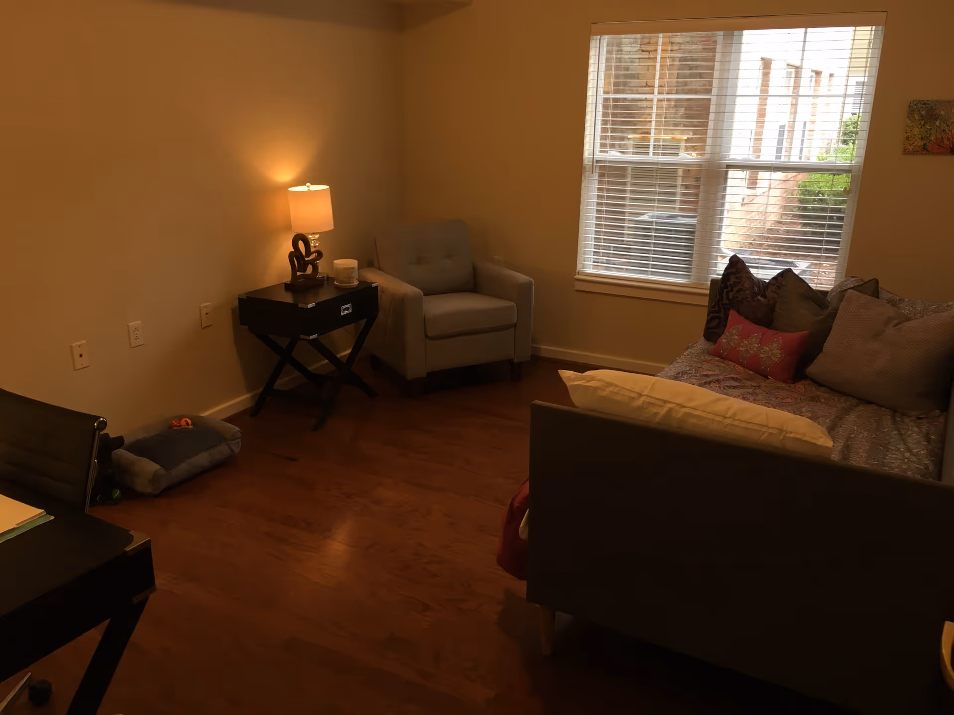 A cozy living room with wooden flooring, a gray armchair next to a black side table with a lamp and decorative item, a couch with multiple pillows, and a window with white blinds letting in natural light.