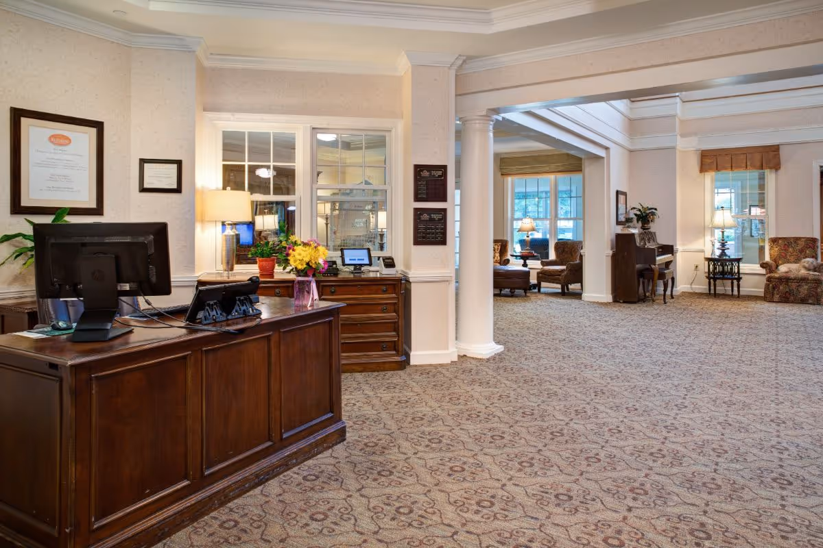 A spacious and well-lit reception area in an assisted living facility featuring a wooden reception desk with a computer, phone, and a vase of flowers. Behind the desk are windows and a lamp on a wooden cabinet. The room has patterned carpet, white columns, and comfortable seating areas with armchairs and lamps in the background.