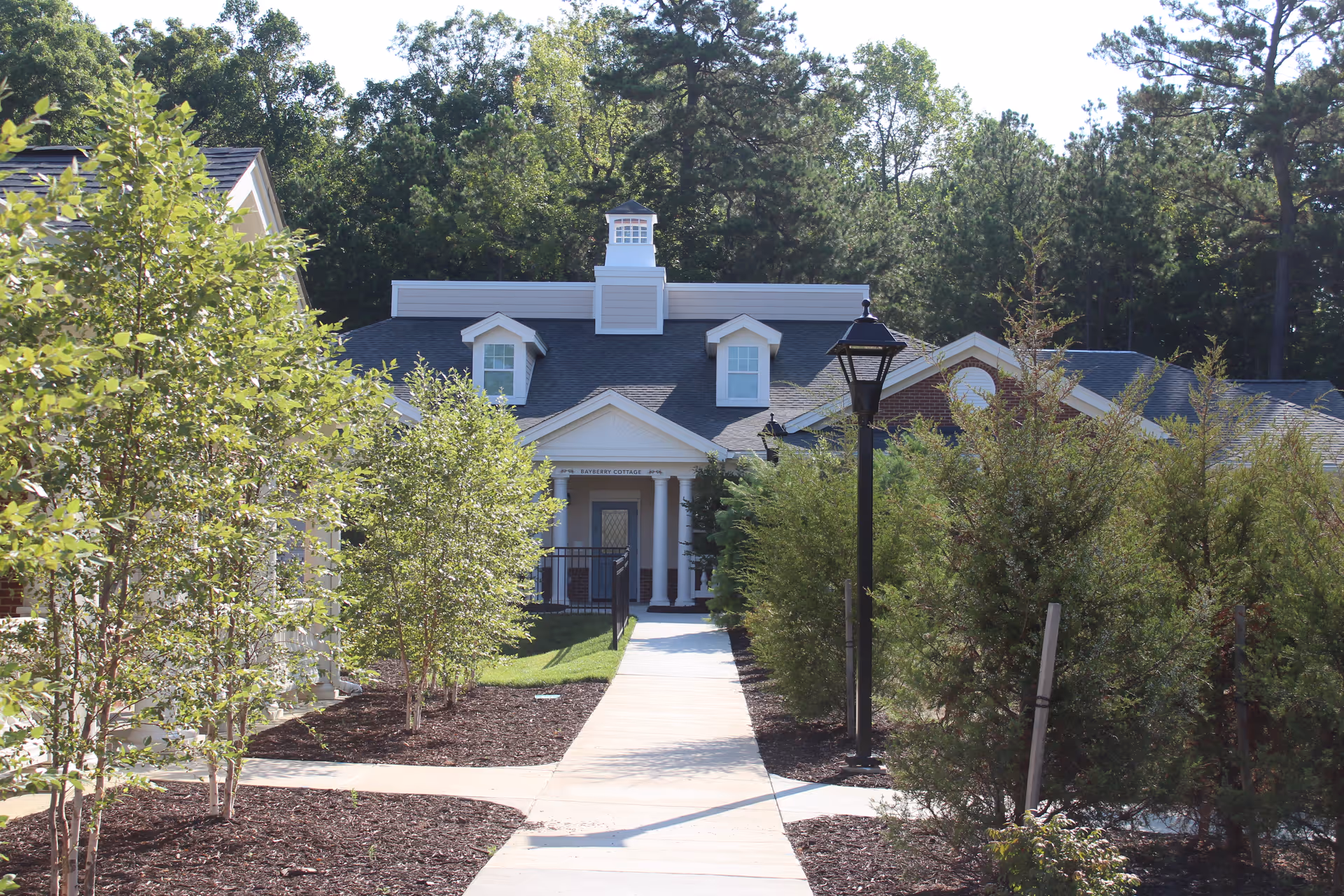 A paved walkway leads through landscaped greenery with small trees and bushes toward a single-story building with a gray roof, white columns, and dormer windows, surrounded by tall trees in the background.