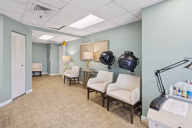Interior view of a salon area in a senior living facility with light green walls, beige carpet, and white ceiling tiles. The room features four white cushioned chairs, two black hair dryers on stands, two table lamps on a glass and metal table, framed artwork on the wall, and a manicure station with various nail care products and a lamp.