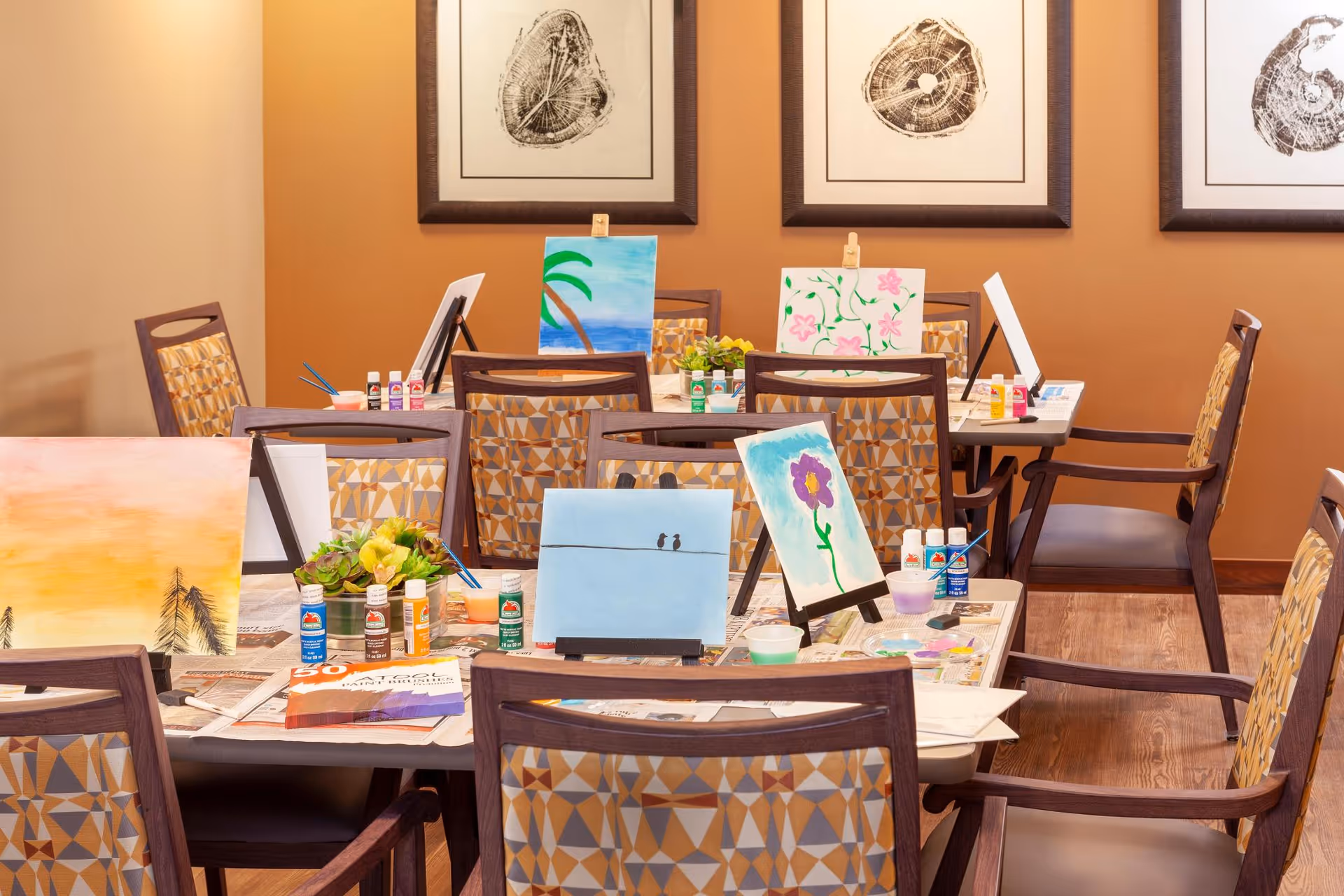 A communal interior room with tables and chairs set up for a painting activity, showing canvases, paint bottles and brushes.