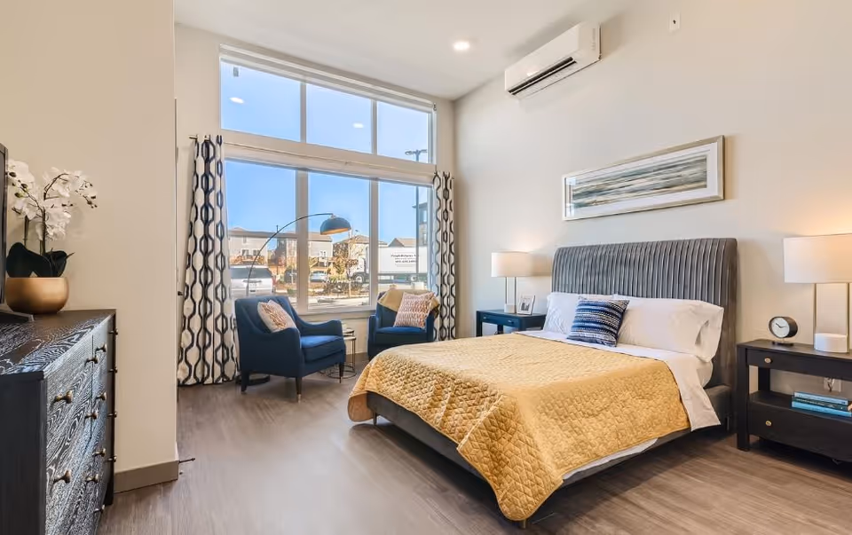 A bright and modern bedroom with a large window letting in natural light. The room features a bed with a yellow quilt and blue and white pillows, two dark blue armchairs with patterned cushions near the window, a dark wooden dresser with a potted white orchid, two nightstands with lamps and a clock, and a piece of abstract art hanging above the bed.