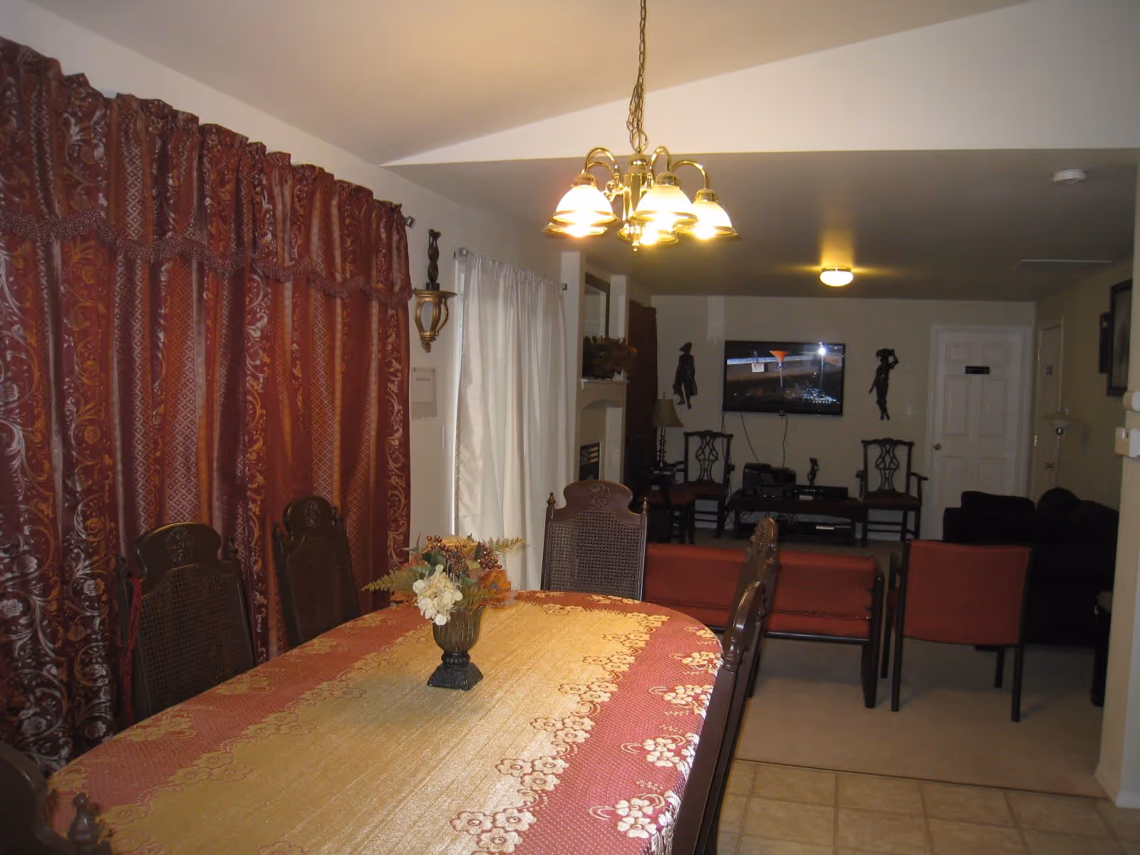 Interior view of a dining area with a long table covered by a red and gold patterned tablecloth and six wooden chairs. A floral centerpiece is on the table. To the left, there are red patterned curtains and white sheer curtains. In the background, there is a living room area with red chairs, a TV mounted on the wall, and decorative wall hangings.