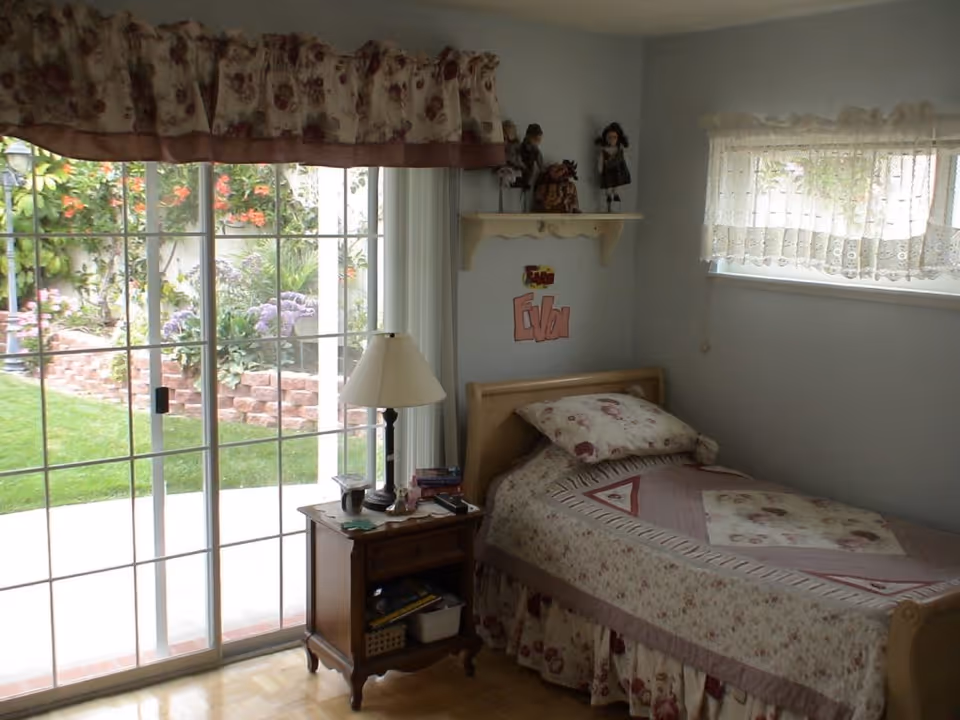 A small bedroom with a single bed covered in a floral quilt and matching pillow. Next to the bed is a wooden nightstand with a lamp, books, and other small items. Above the bed is a small wall shelf holding decorative dolls. The room has a large sliding glass door with floral curtains that opens to a garden with green grass and flowering plants. There is also a small window with lace curtains on the adjacent wall.