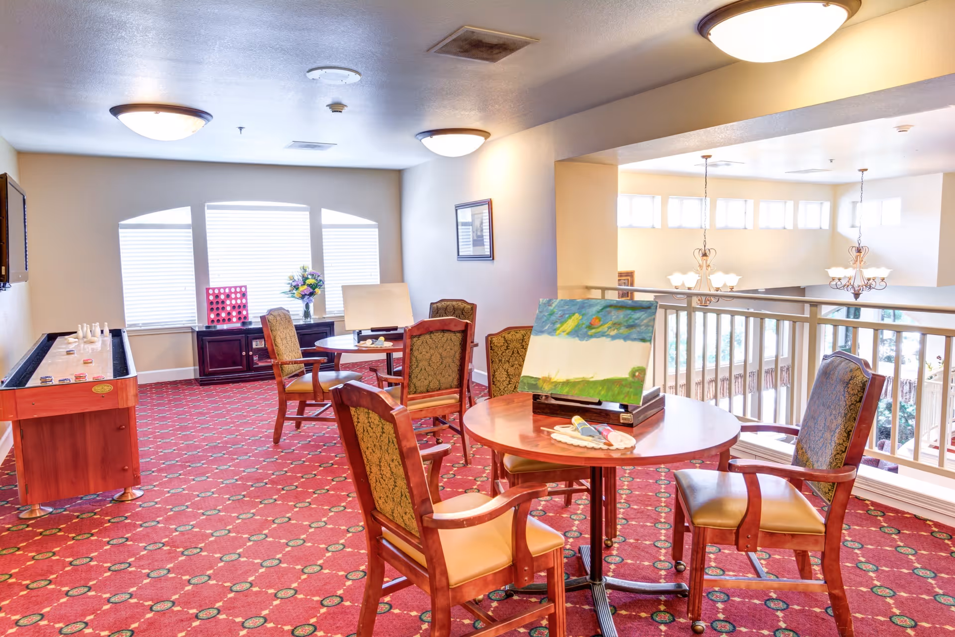 A bright common area with red patterned carpet, several wooden chairs with cushioned seats around circular tables, and a shuffleboard table along the left wall. One table has a painting and art supplies on it. Large windows with blinds let in natural light, and chandeliers hang from the ceiling in the adjacent open space.